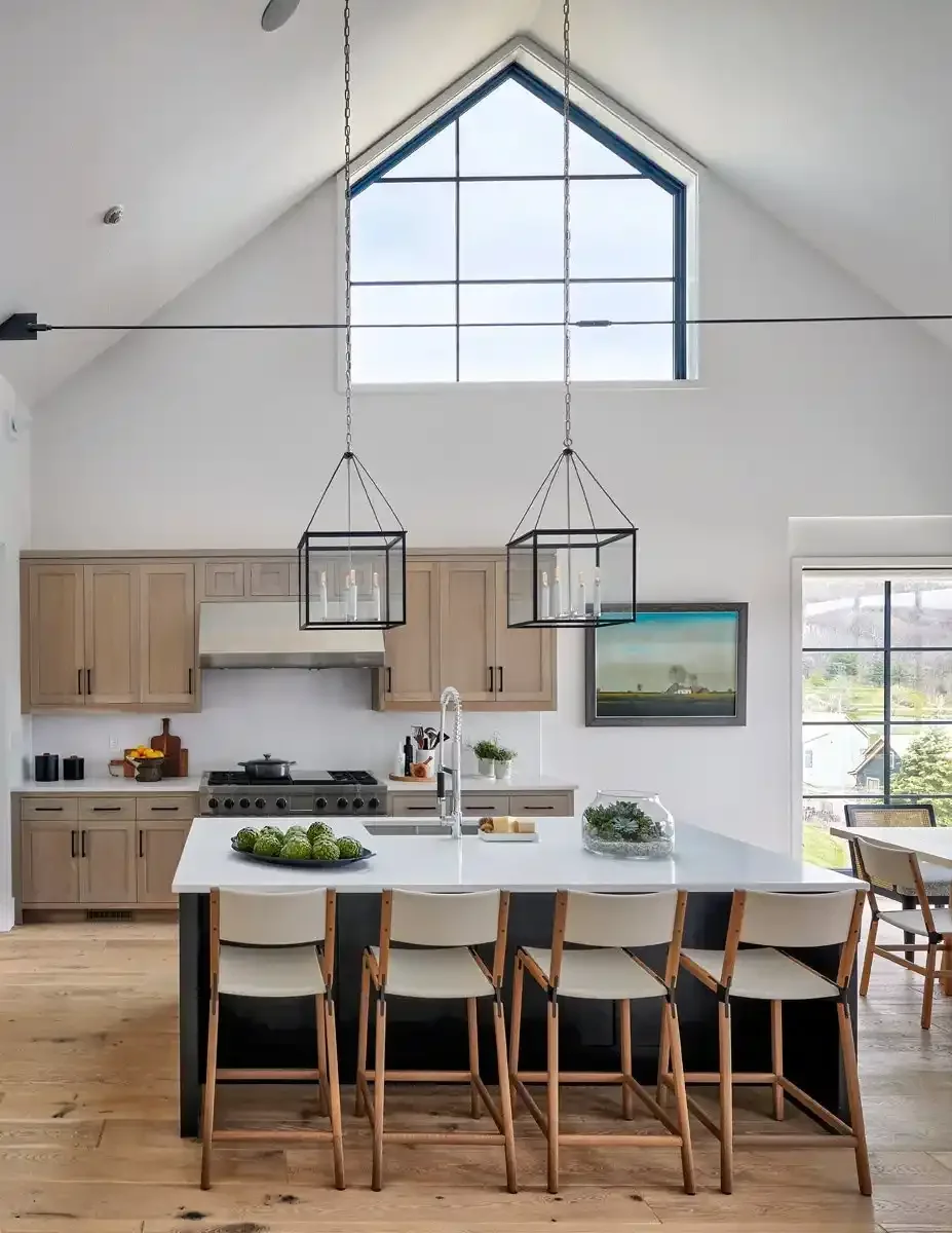 Modern kitchen with a white island, light wood cabinets, and two black pendant lights hanging from a high vaulted ceiling with a large triangular window.