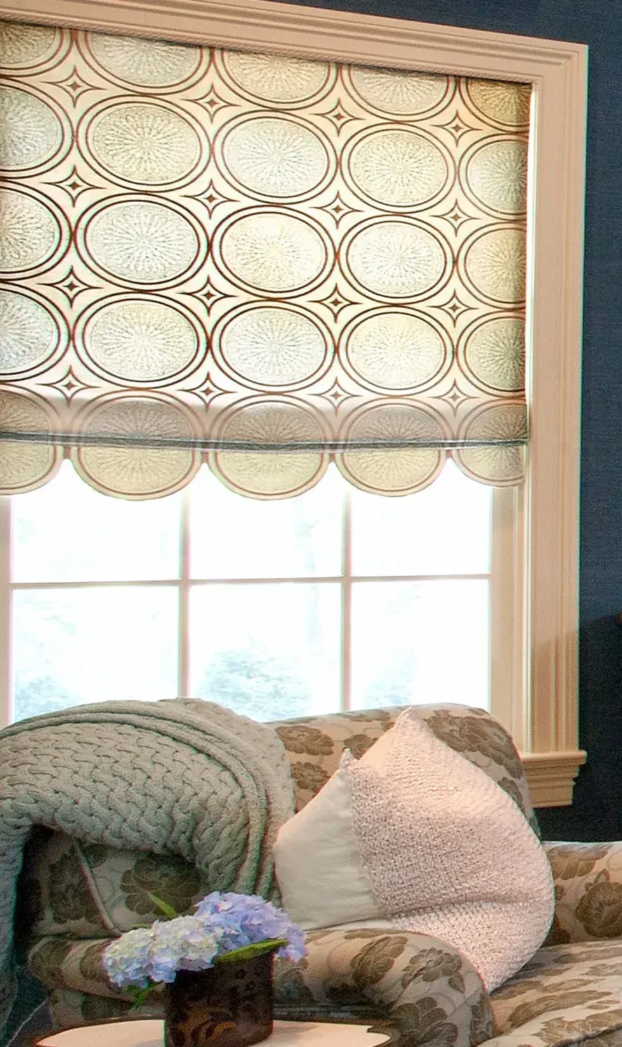 A window with patterned blinds, filled with natural light, sitting above a floral-patterned sofa with a beige knitted pillow and a white pillow. A small table in front holds a flower arrangement.