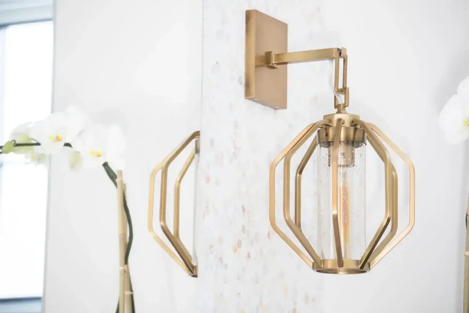 Modern gold chandelier with geometric design hanging on white wall, partially visible orchid plant on the left.