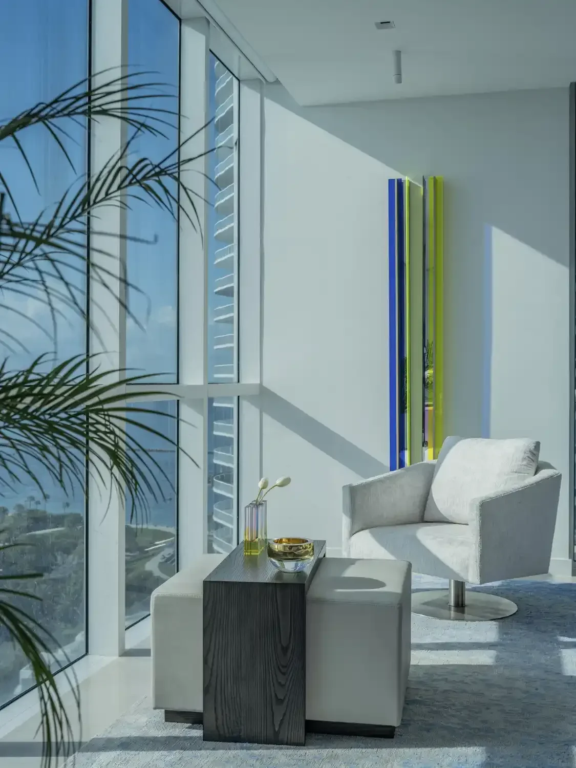 Modern living room corner with white armchair, white ottoman, wooden coffee table, decorative vases, colorful vertical art sculpture, large windows with city view, sunlight casting shadows, indoor plant.