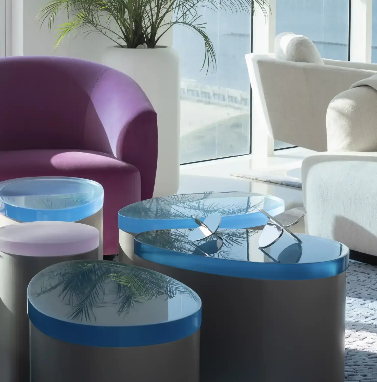 Modern indoor space with colorful seating and glass-top tables, large potted plant, and floor-to-ceiling window showing ocean view.