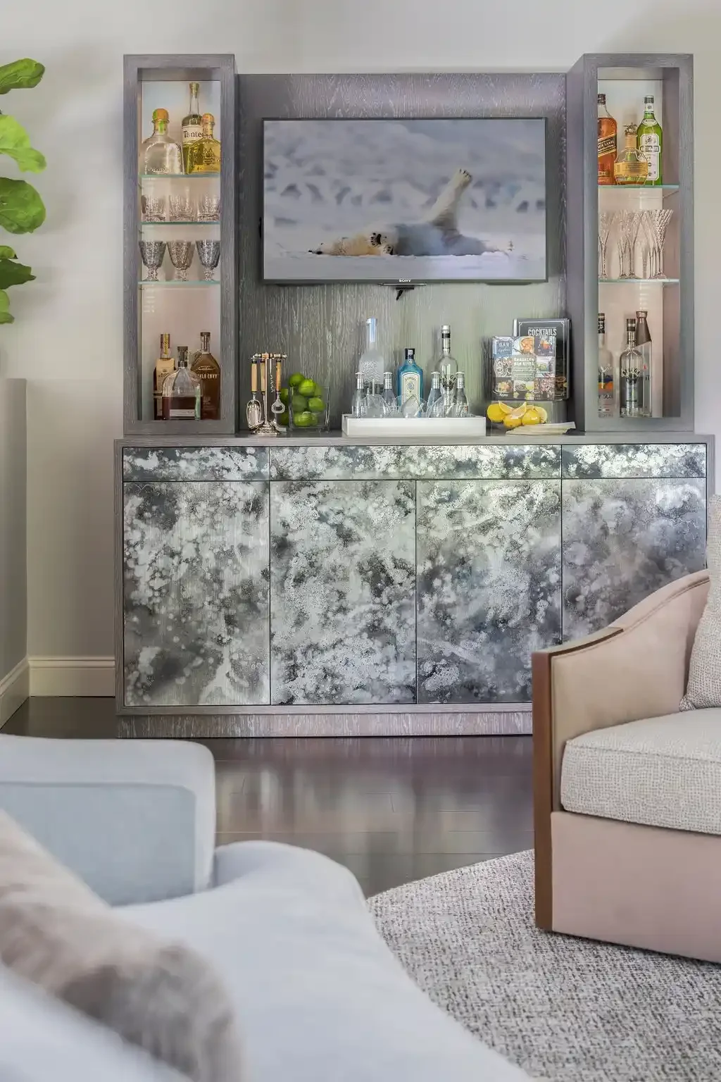 Modern bar area with a mounted TV showing a polar bear, surrounded by glass cabinets with liquor bottles and glassware, and a textured cabinet front with a speckled pattern.