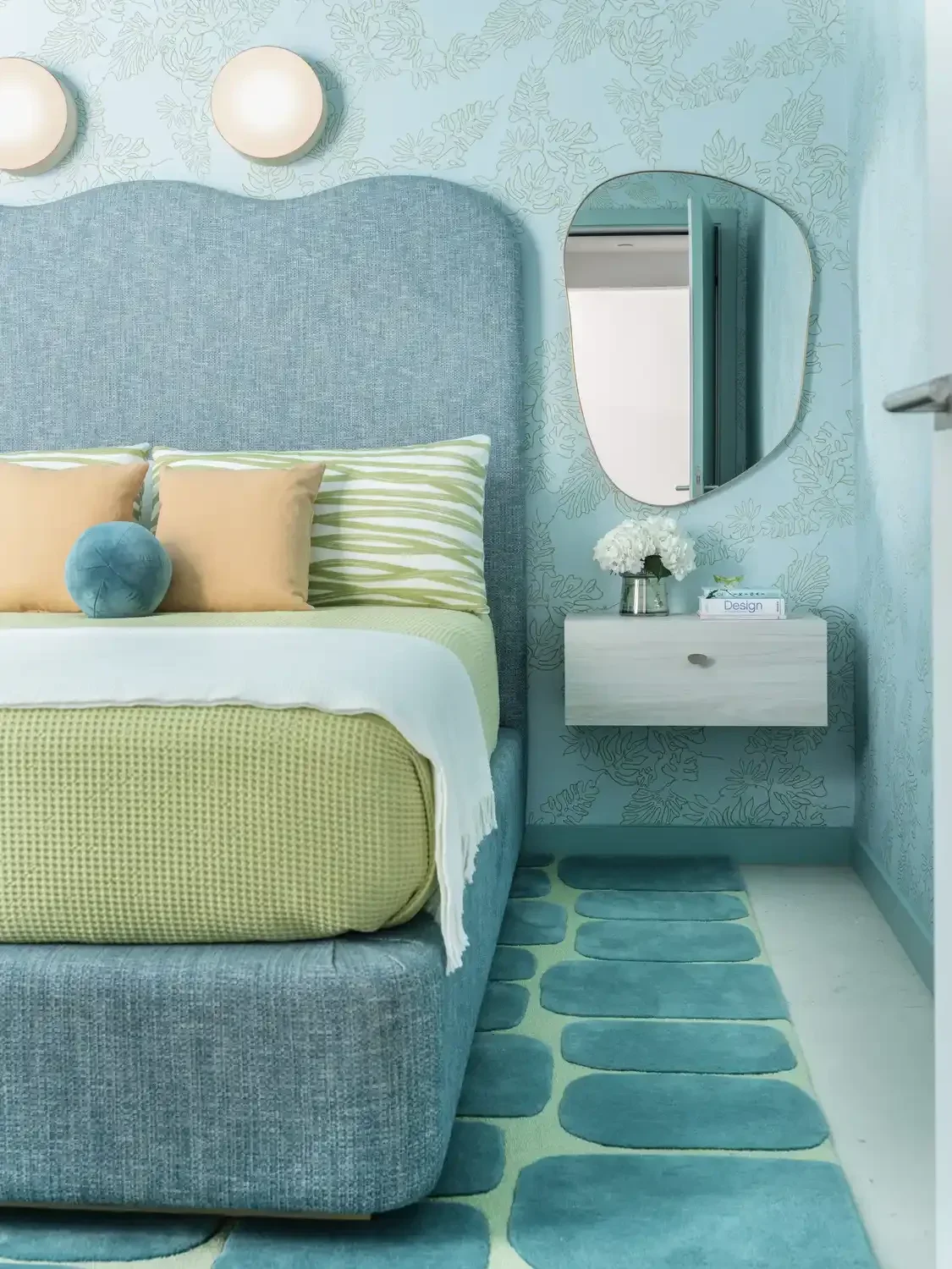 A bedroom with a blue upholstered bed, yellow and green pillows, a white nightstand with a bouquet of white flowers, a round mirror on a blue floral-patterned wall, and a blue and green oval-striped rug.