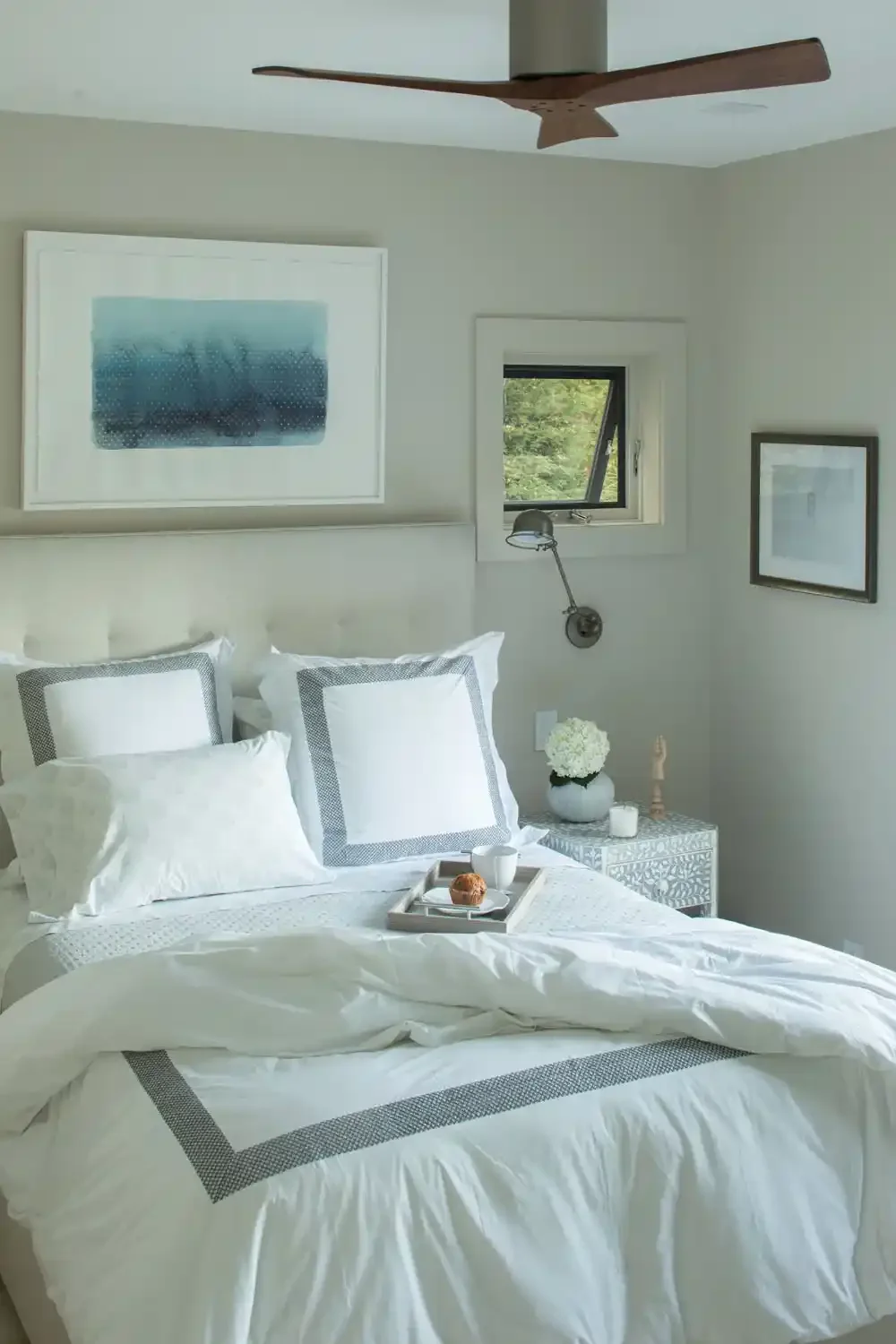A cozy bedroom with a neatly made bed, white bedding with gray accents, a tray with a cup, pastry, and a white mug on top. There is a bedside table with a vase of white flowers, a candle, and a small decorative item. The room has artwork on the walls