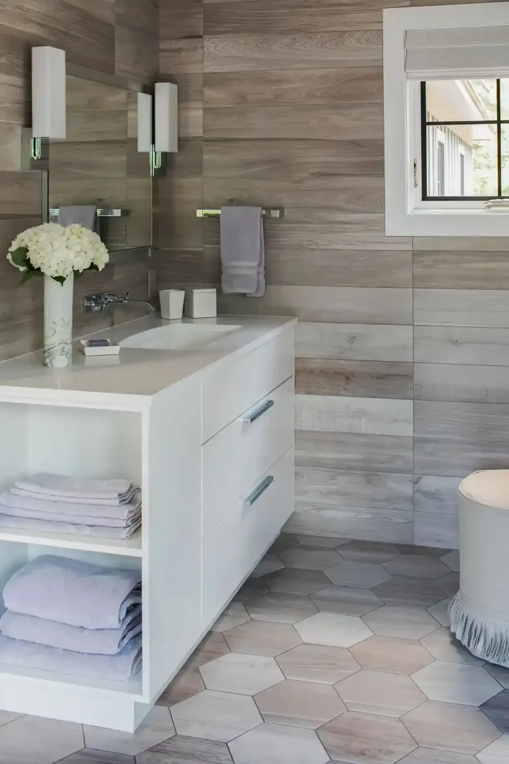 A modern bathroom with a white vanity, a vase of white flowers, folded towels, a small soap dish, two cups, gray towels hanging, a mirror, wall lights, a window, and hexagon floor tiles.