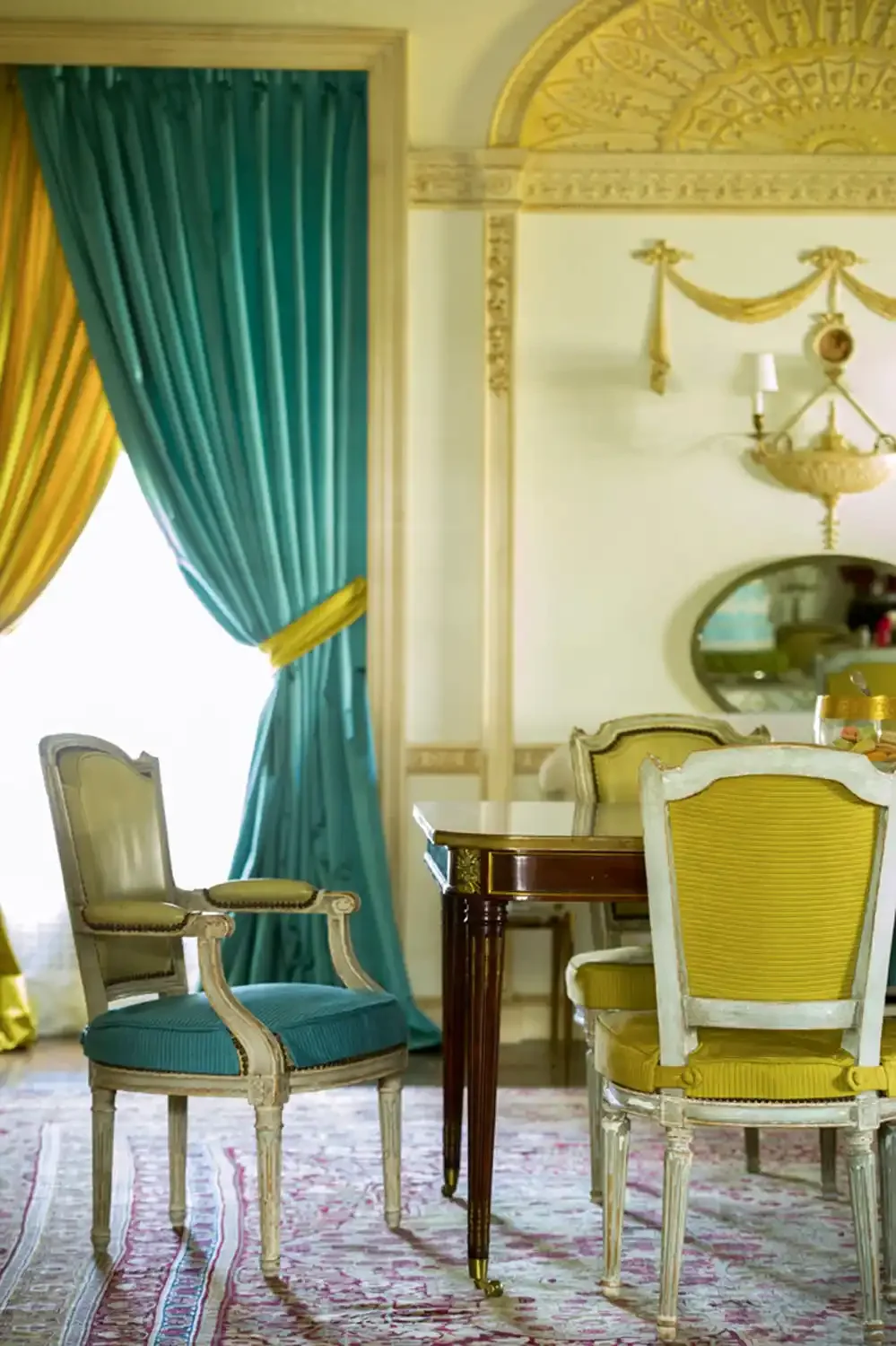 Elegant room with yellow and teal curtains, vintage chairs with yellow and teal upholstery, a dark wood table, a large ornate mirror, and a chandelier with gold detailing.