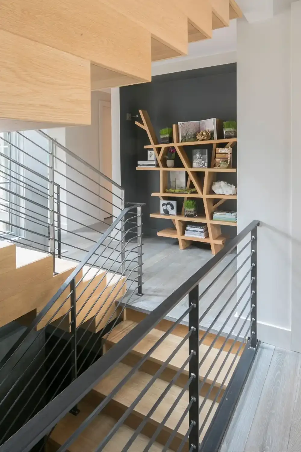 Modern interior staircase with metal railing and wooden steps, overlooking a small accent wall with a wooden bookshelf and decorative items.