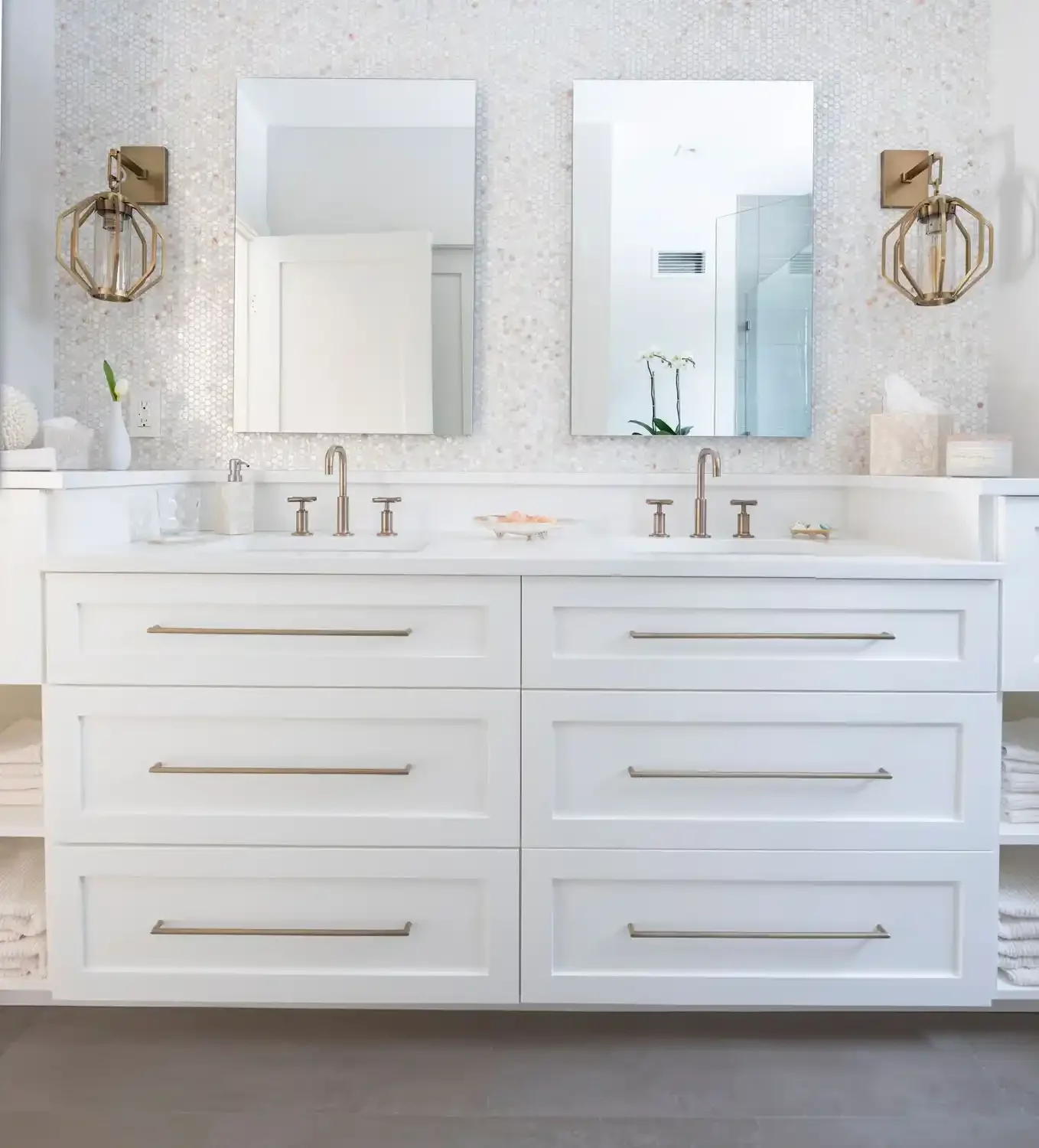 A modern white bathroom vanity with six large drawers with gold handles, two undermount sinks with gold faucets, and two wall-mounted gold light fixtures with glass shades. Two mirrors are mounted above the sinks, reflecting a bright, minimalist spac
