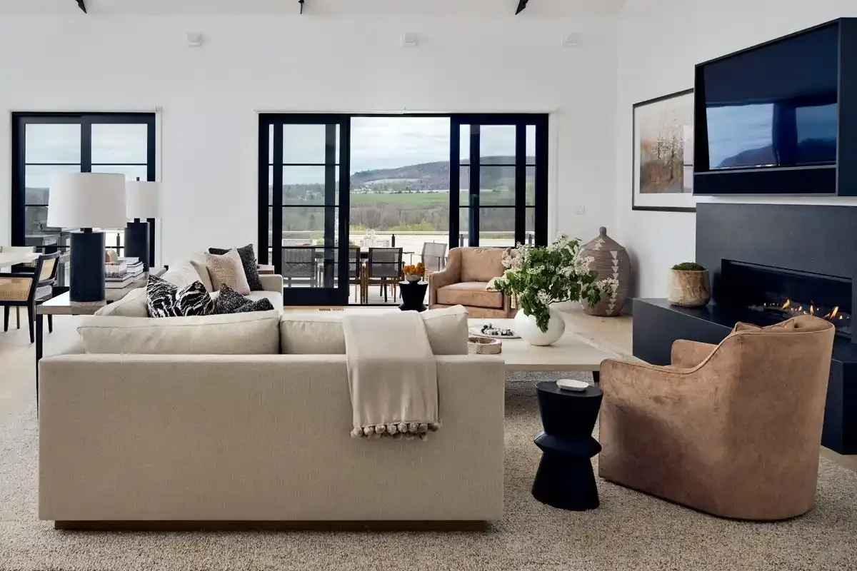 Modern living room with beige sofa, tan armchair, and black side table, with a fireplace and TV on the wall, large windows overlooking a scenic outdoor view, and decorative plants and artwork.