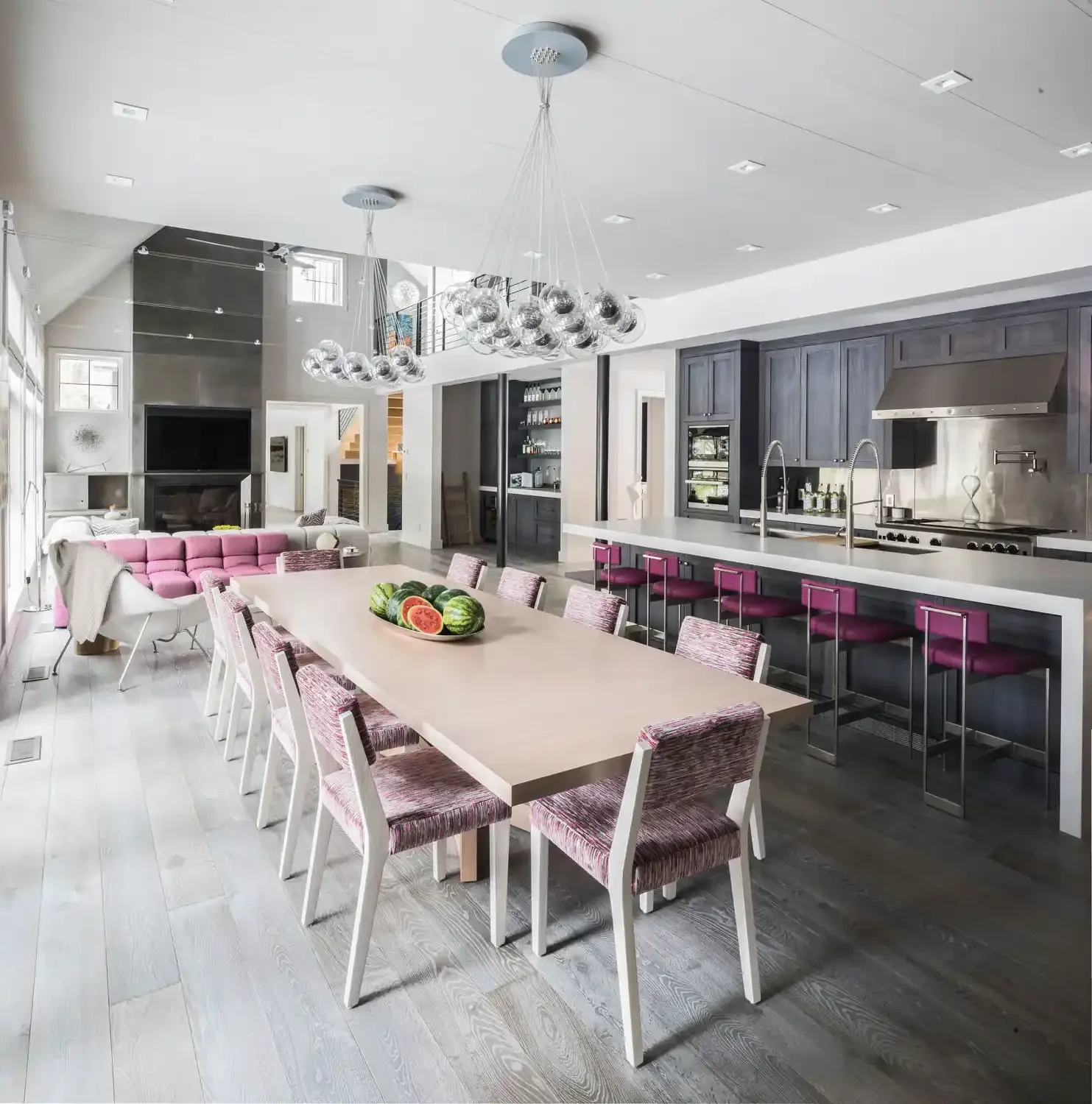 Open-concept kitchen and dining area with a large white dining table, pink upholstered chairs, a modern kitchen with dark gray cabinets, a stainless steel range hood, a white island counter with pink bar stools, a pink striped sofa, and a chandelier 