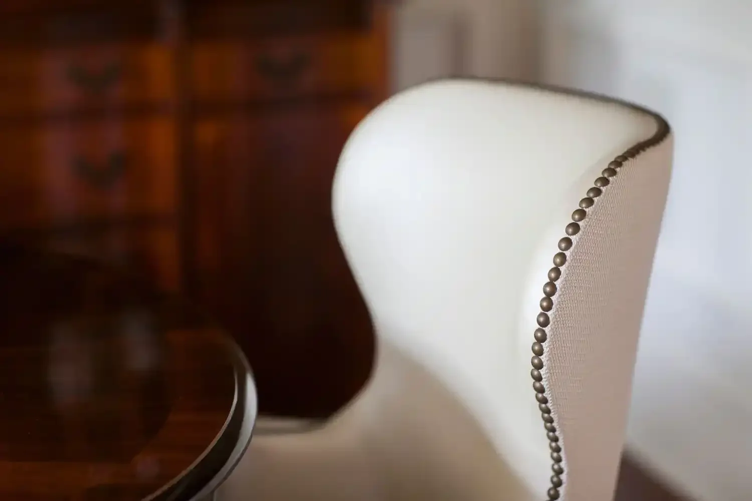 Close-up of a white upholstered dining chair with decorative nailhead trim along the edge, next to a dark wooden dining table, with a blurred wooden cabinet in the background.
