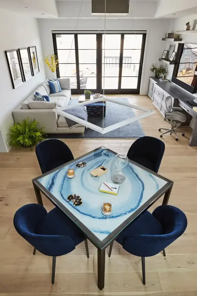 A modern open-plan living room and dining area with a glass-topped table and four blue chairs in the foreground. The living room features a white sofa with blue and gray pillows, a gray area rug, a black coffee table, and a wall of large windows with