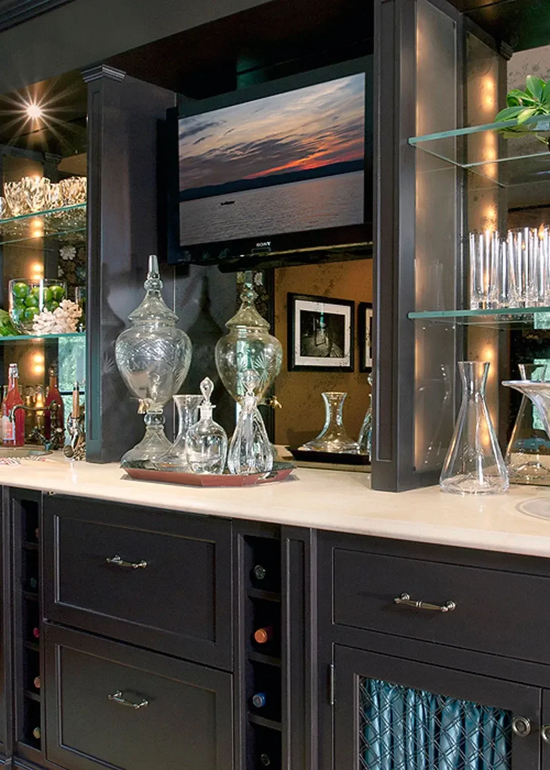 A home bar area with dark cabinetry, glass decanters, and a television showing a sunset over water.