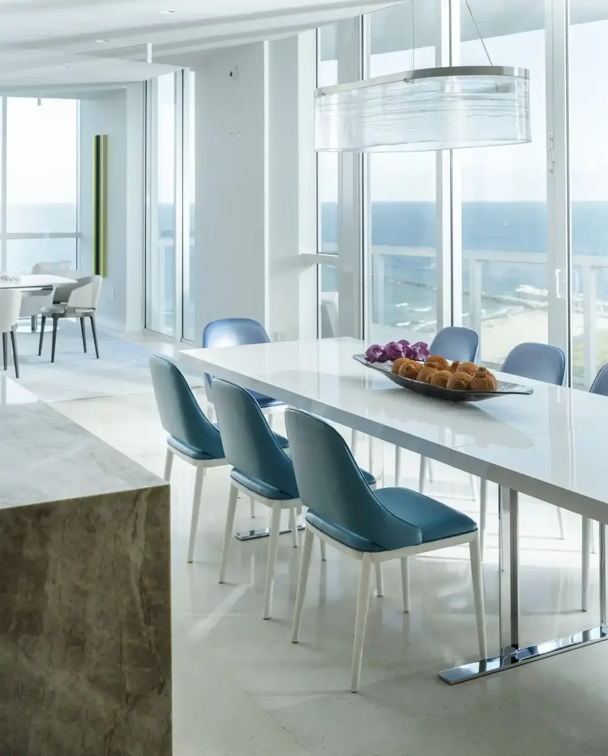 Modern dining area with large windows, a white table, teal chairs, and a decorative celling light fixture. A black tray with purple and orange pastries is on the table.