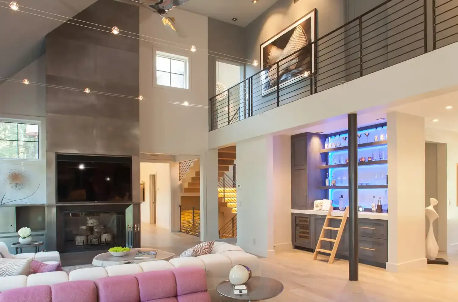 Open-concept living room with a two-story ceiling, large windows, a modern fireplace, a flat-screen TV, a pink sectional sofa, a coffee table with a decorative bowl, a bar area with blue lighting, and a staircase with wooden steps and black railings.
