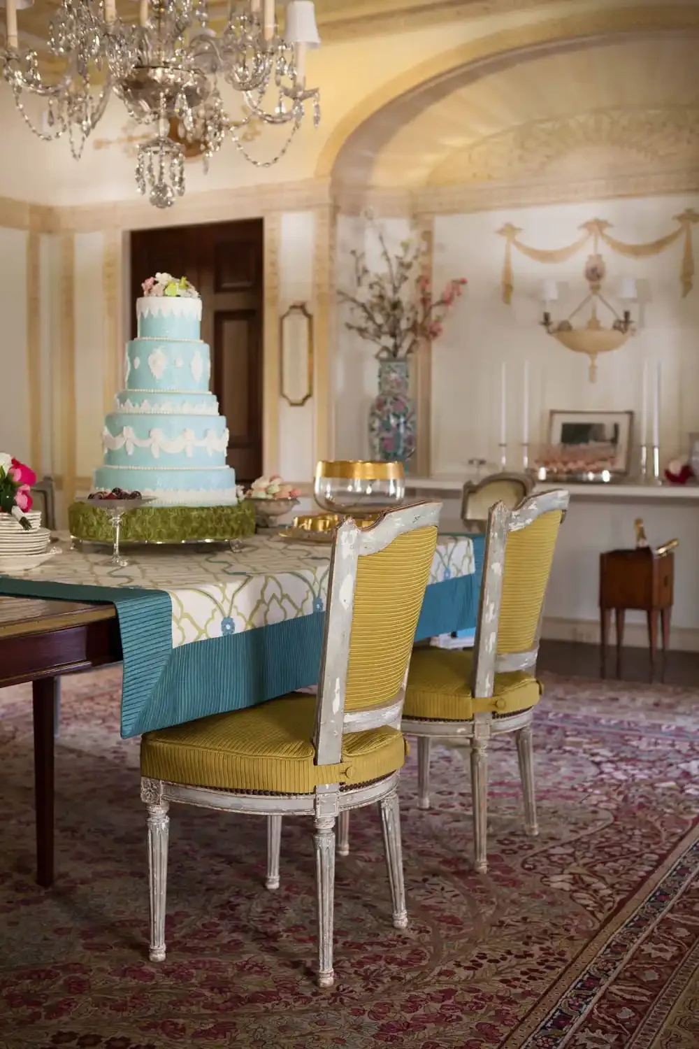 Elegant dining room with ornate chandelier, a layered turquoise and white cake on the table, floral decorations, and vintage chairs.
