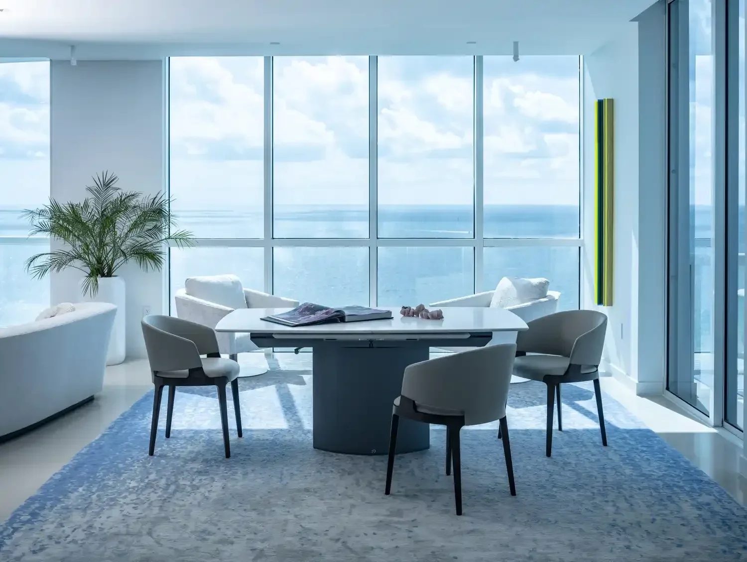 Modern dining area with a round table, four chairs, white armchairs, a large potted plant, large windows with ocean view, and a blue patterned rug.