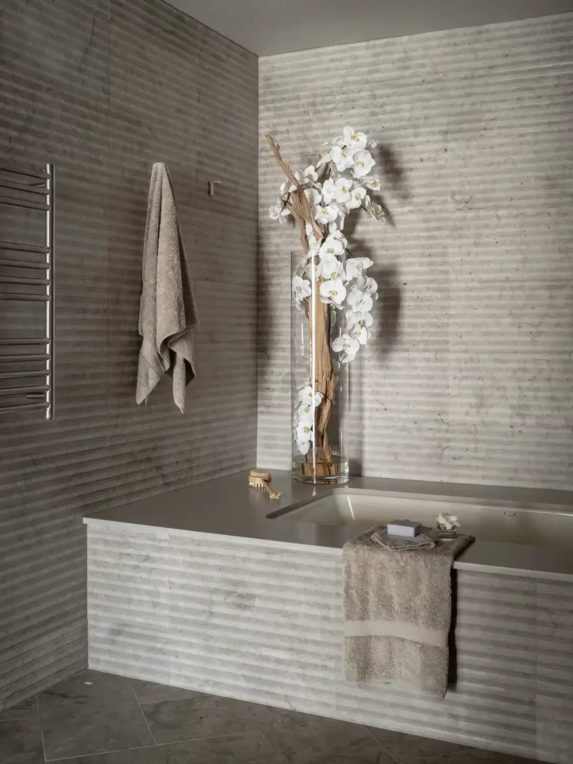 Bathroom with textured gray tiles, a towel hanging on a rack, a large glass vase with white orchids and wooden branches, a bathtub with a beige towel, soap, and bath accessories.