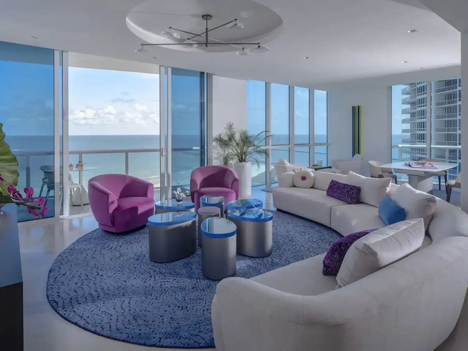 Living room with large glass windows showcasing an ocean view, white sofa with purple and blue pillows, two purple armchairs, and modern side tables, with a blue patterned rug and tall plants.
