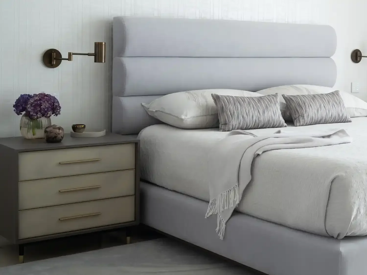 Bedroom with a bed, beige and gray bedding, a gray headboard, and a gray nightstand with a purple flower vase, a small decorative object, and a candle. Wall-mounted lamps are on either side of the headboard.
