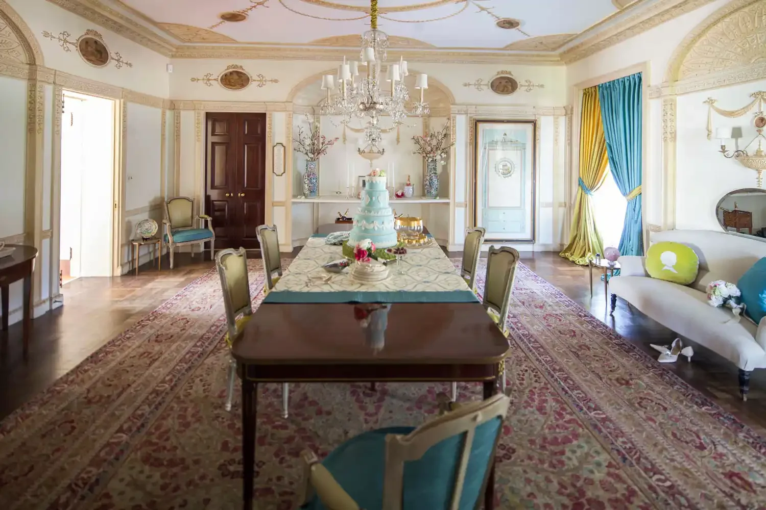 A luxurious dining room with a large table set for celebration, topped with a multi-tiered cake, and surrounded by elegant chairs. The room features ornate molding, a chandelier, colorful curtains, and vintage furniture, with an area rug on the woode