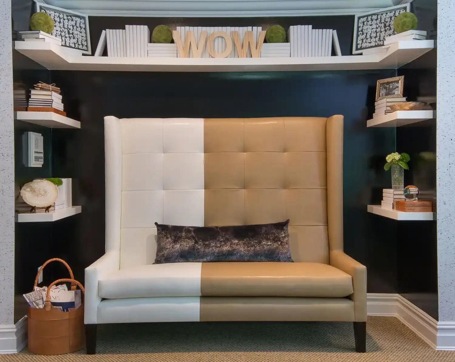 A cozy nook with a large beige and white tufted headboard, decorated with a dark pillow. Above the headboard is a wall with floating shelves holding books, decorative objects, and greenery. The top shelf has wooden 'WOW' letters and green moss balls.