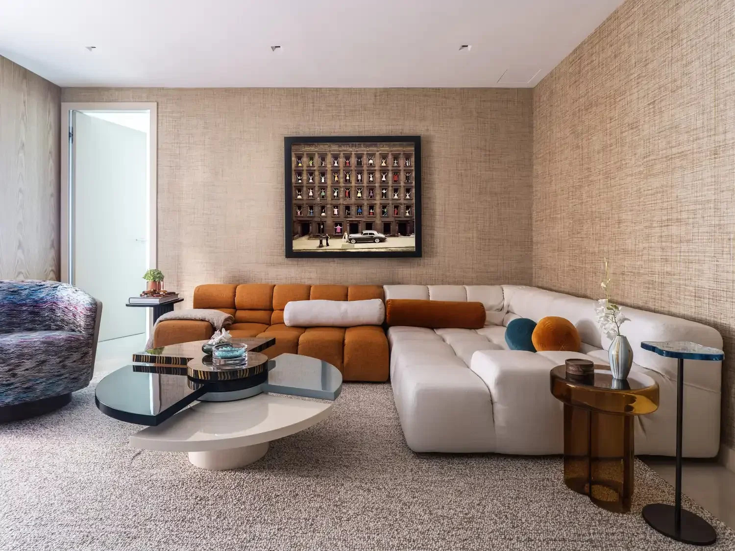 Modern living room featuring a sectional sofa with white, orange, and brown cushions, a textured wall with a framed photo, a multicolored armchair, and a set of stylish coffee tables with decorative items and a vase of white flowers.