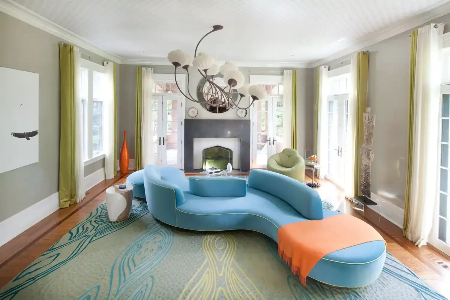 Bright living room with large windows, green curtains, a central fireplace, a unique blue curved sofa with an orange throw blanket, a green armchair, and modern decor including a tall orange vase and a sculpture near the window.