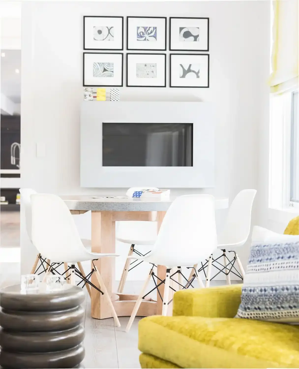 Modern living room with a round dining table surrounded by white chairs, a wall-mounted fireplace, a gallery wall with black-framed abstract art, and a yellow sofa.
