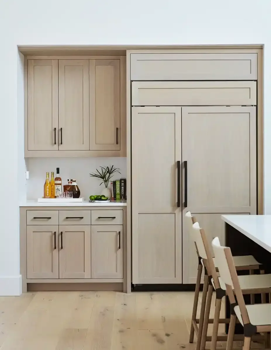 Kitchen with built-in wooden cabinets, a white countertop, a large black refrigerator, a small bar area with bottles and glasses, potted plant, and wooden chairs.