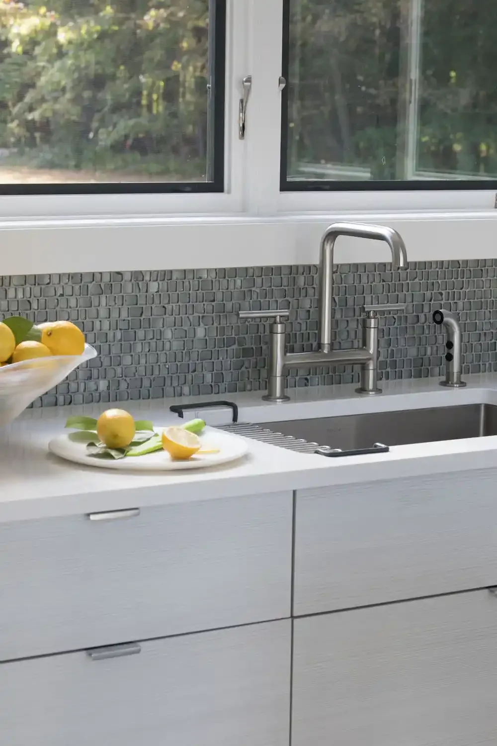 Kitchen sink with a modern faucet, a gray tiled backsplash, a window with a view of trees, a white counter with a lemon and a lemon half, and a bowl of lemons.