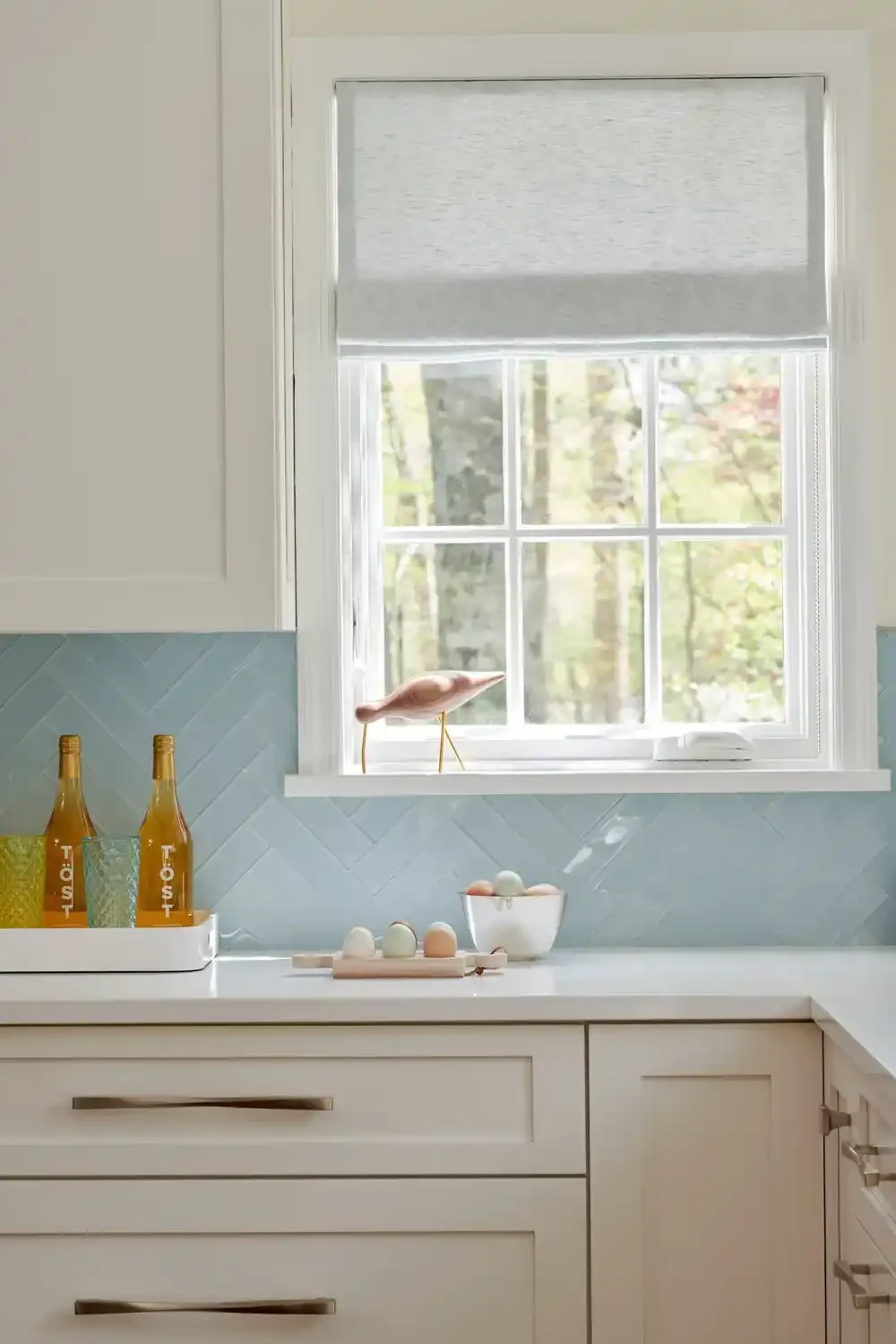 A kitchen countertop near a window with a light blue tile backsplash, two bottles labeled 'TOST', a glass with a yellow-green pattern, a white bowl with pastel-colored eggs, and a whimsical pink bird figurine with gold legs on the windowsill.