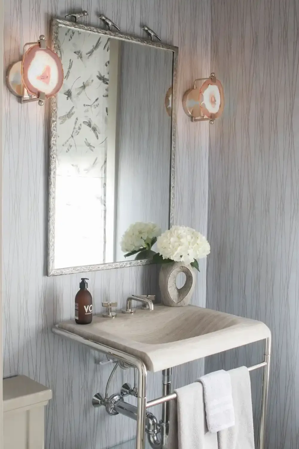 A bathroom with a mirror, two wall-mounted lights, a sink with a flower vase, and a towel hanging on a metal towel rack.