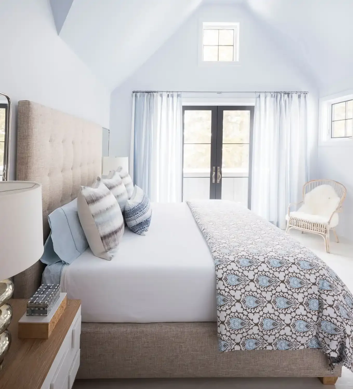 Bright bedroom with white walls, a large bed with striped pillows, a patterned throw blanket, and a tufted headboard. There are windows with white curtains, a wicker chair with a pillow, and a nightstand with decorative items.