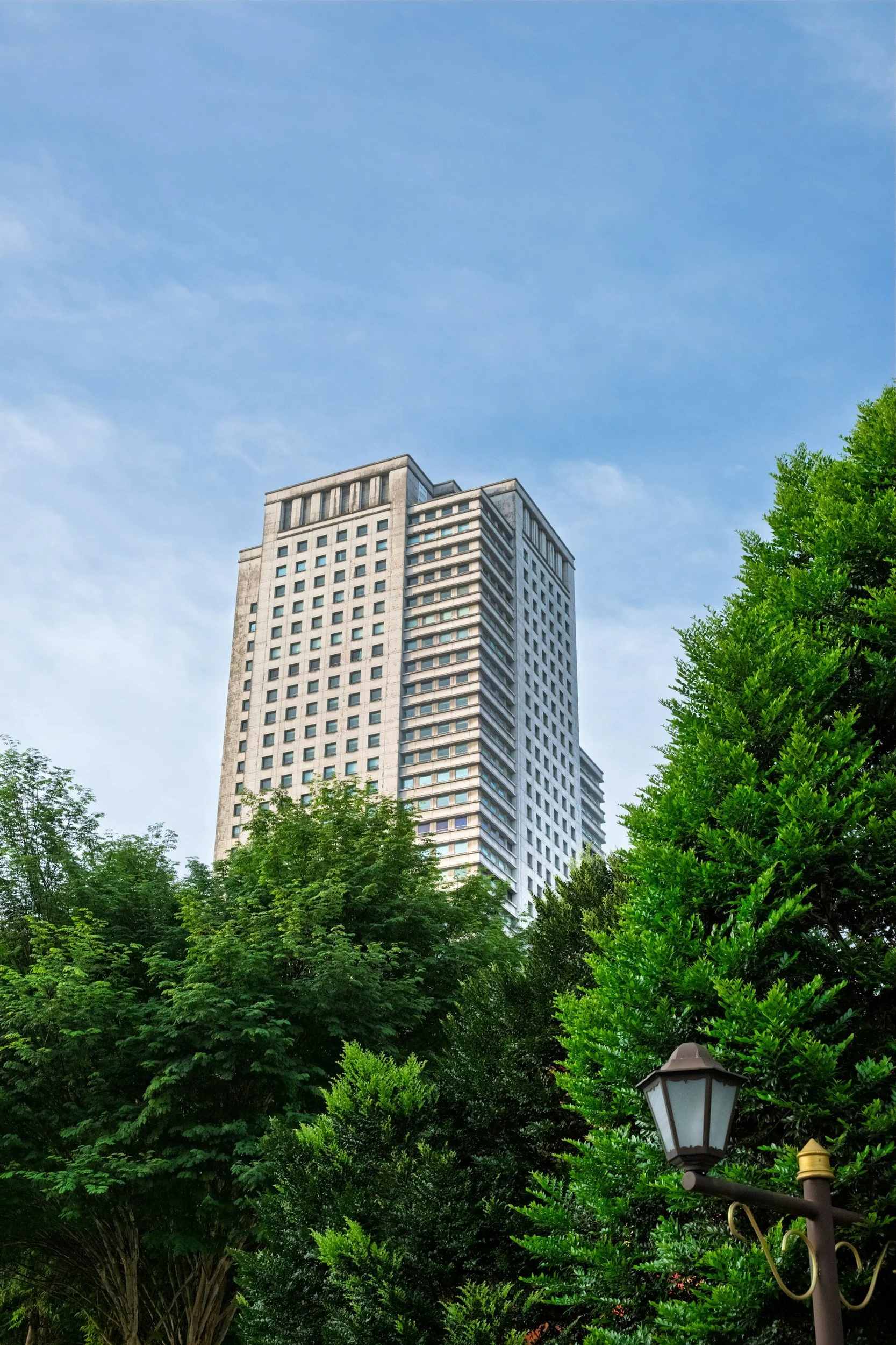 Tall building with many windows rising above green trees and bushes, blue sky with some clouds, and a street lamp in the foreground.
