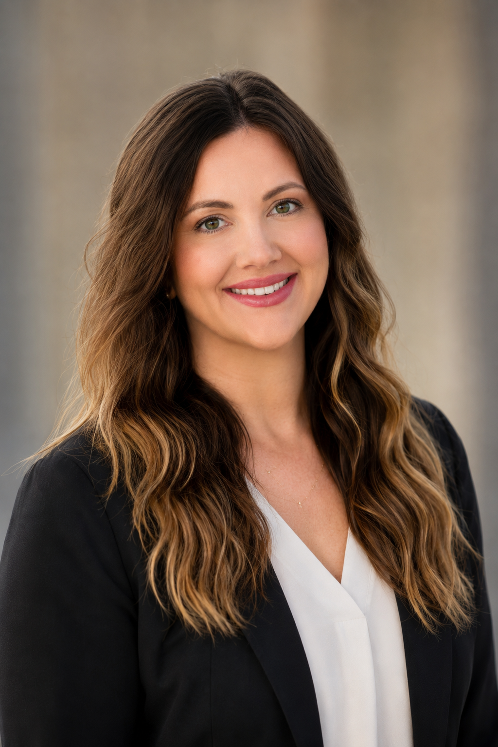 Professional headshot of a woman with long, wavy brown hair, wearing a black blazer and white top, smiling against a neutral background.