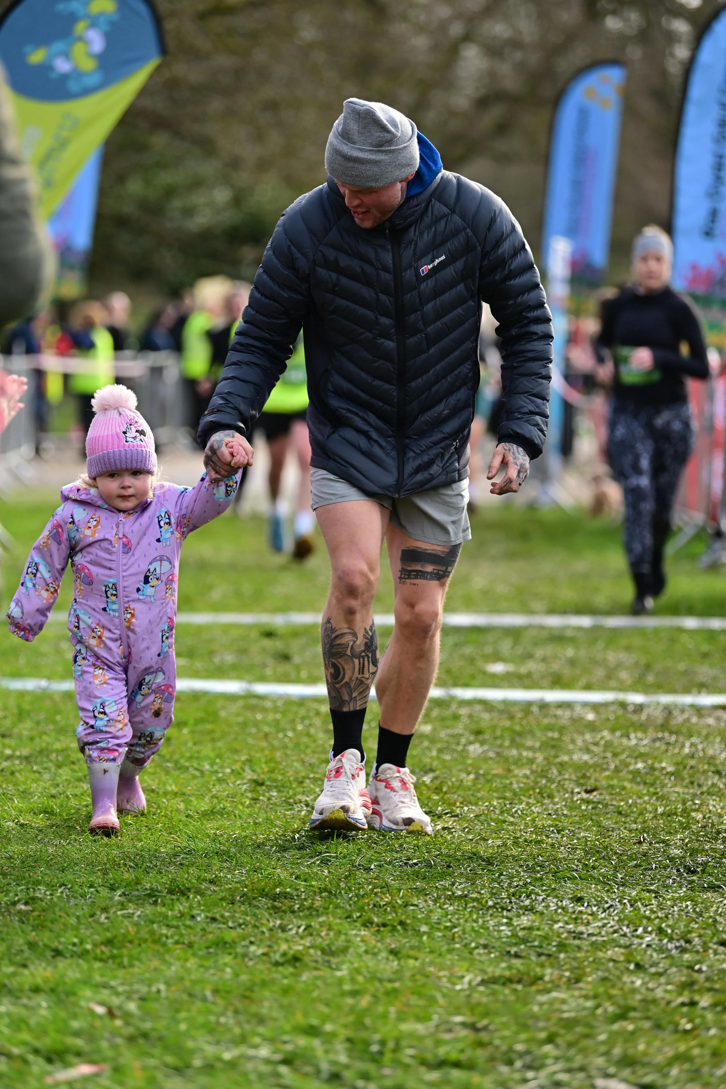A man and a young girl holding hands walking on a grassy field during a running event. The man is wearing a gray beanie, black puffer jacket, and shorts. The girl is dressed in a colorful onesie with cartoon characters and a pink knit hat with a pom-pom. There are several event flags in the background and other participants.