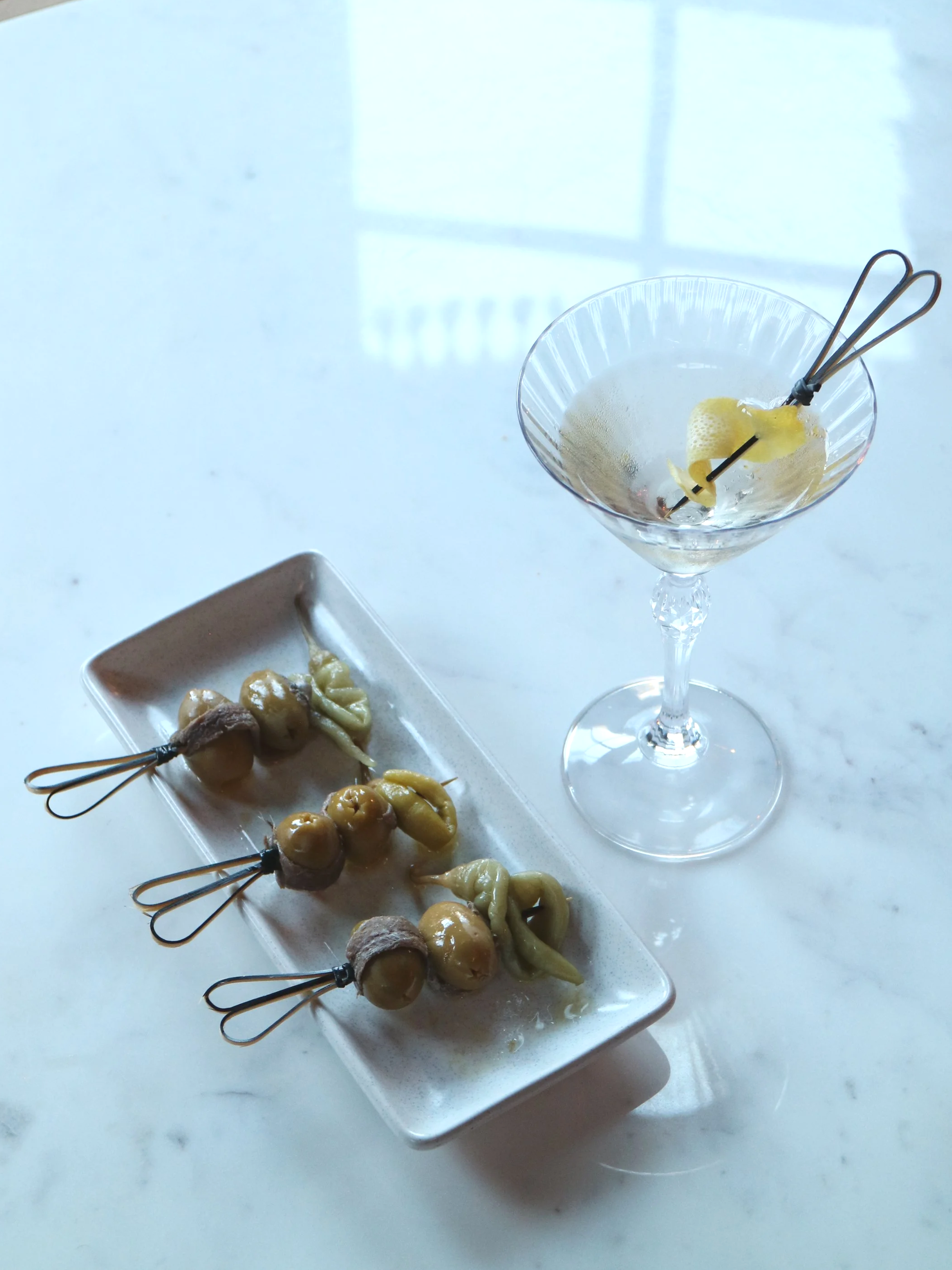 A white rectangular plate with several canapes, and a martini with a lemon twist and cocktail pick, on a white marble surface.