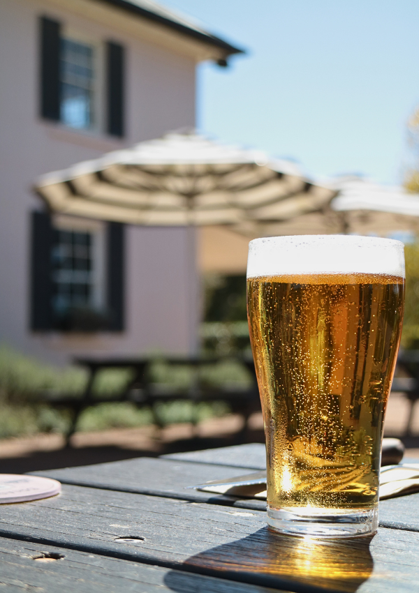 A glass of beer on a wooden table outdoors on a sunny day at a patio with an umbrella and a pub in the background.