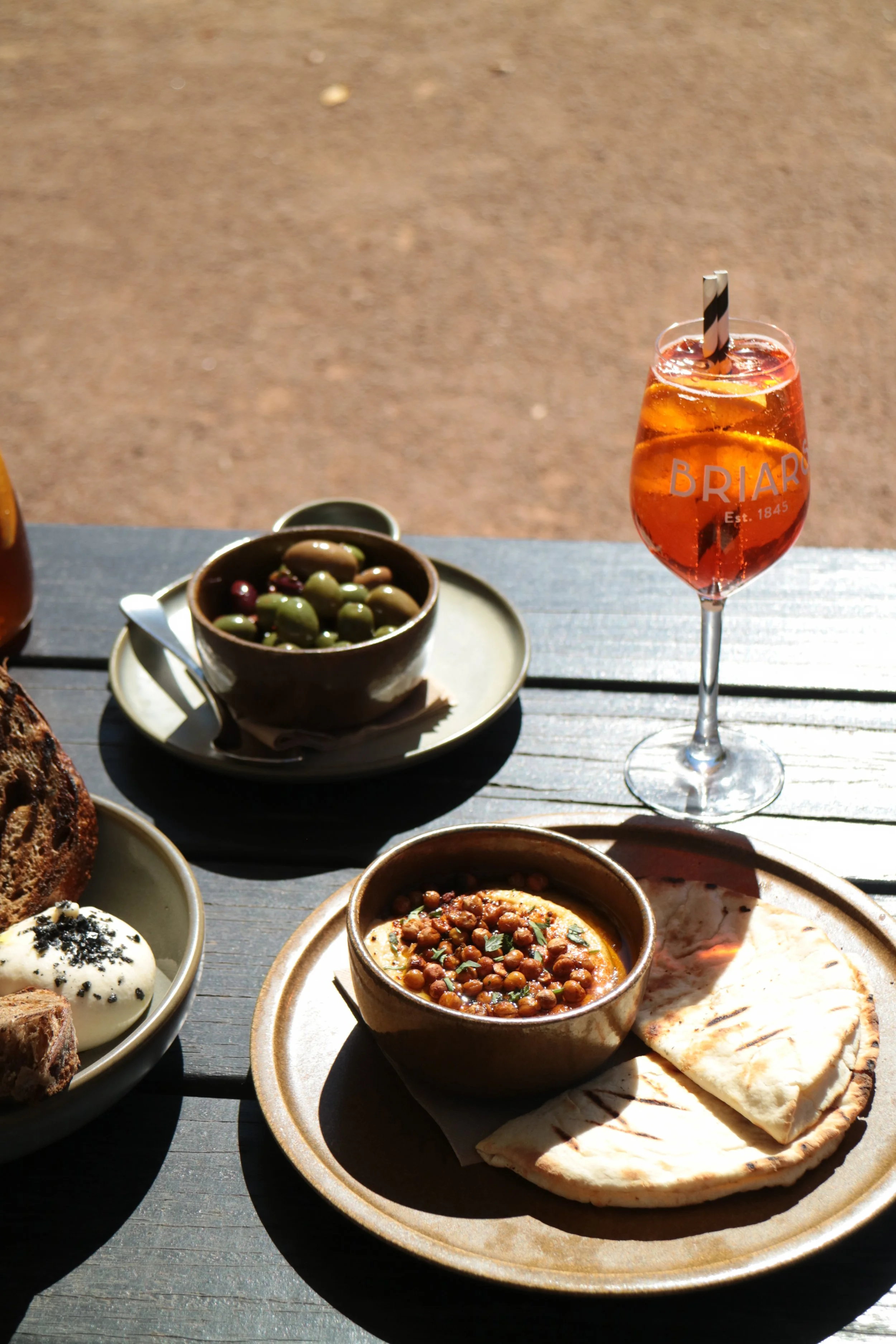A dining table with various dishes and drinks including a bowl of olives, a plate with flatbread and dips, and a glass of aperol with lemon and a straw, outdoors on a sunny day.