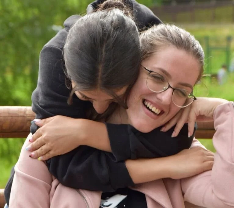 Two women share a joyful hug outdoors, smiling broadly, with a blurred green background.