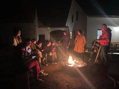 Group of people gathered around a campfire outside at night, some sitting and some standing.