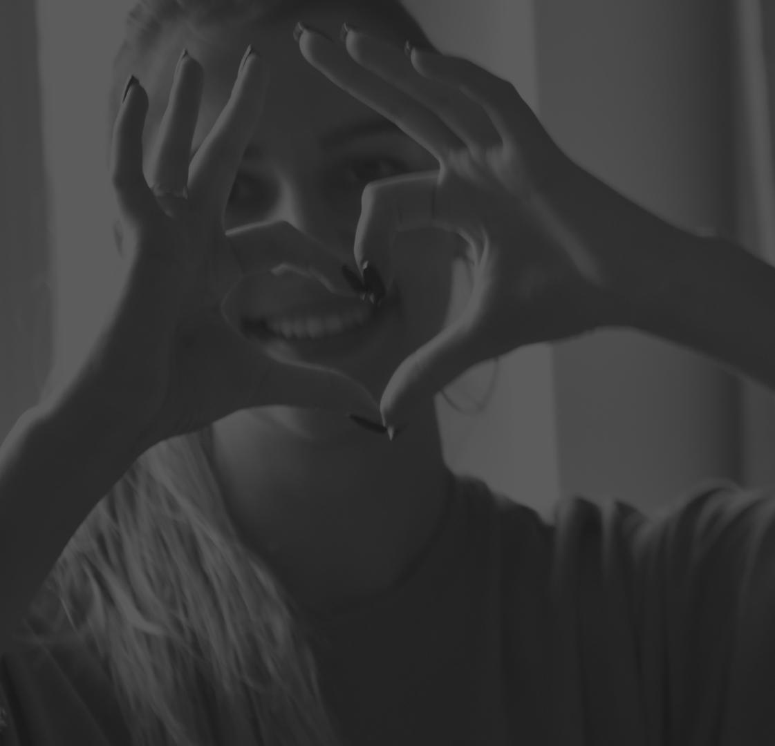 Person making a heart shape with hands, with a face behind it, in black and white.