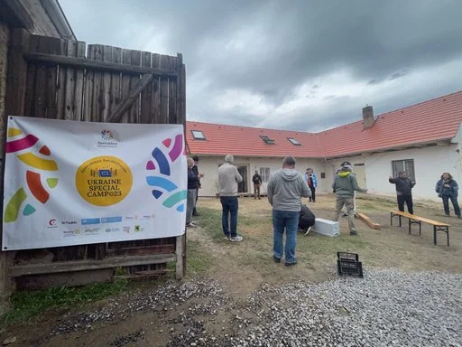 Group of people standing outdoors near a large banner that reads "Ukraine Special Camp" with colorful graphics, in a courtyard area with a wooden fence and a building with a red roof under cloudy skies.