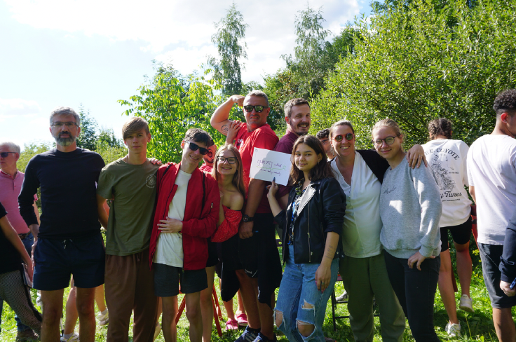Group of young people and adults standing outdoors in a green park, smiling, with trees and blue sky behind them.