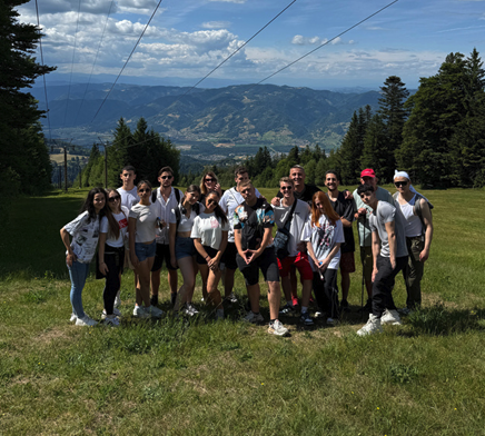 Group of young people standing outdoors on a grassy field with mountains and trees in the background.