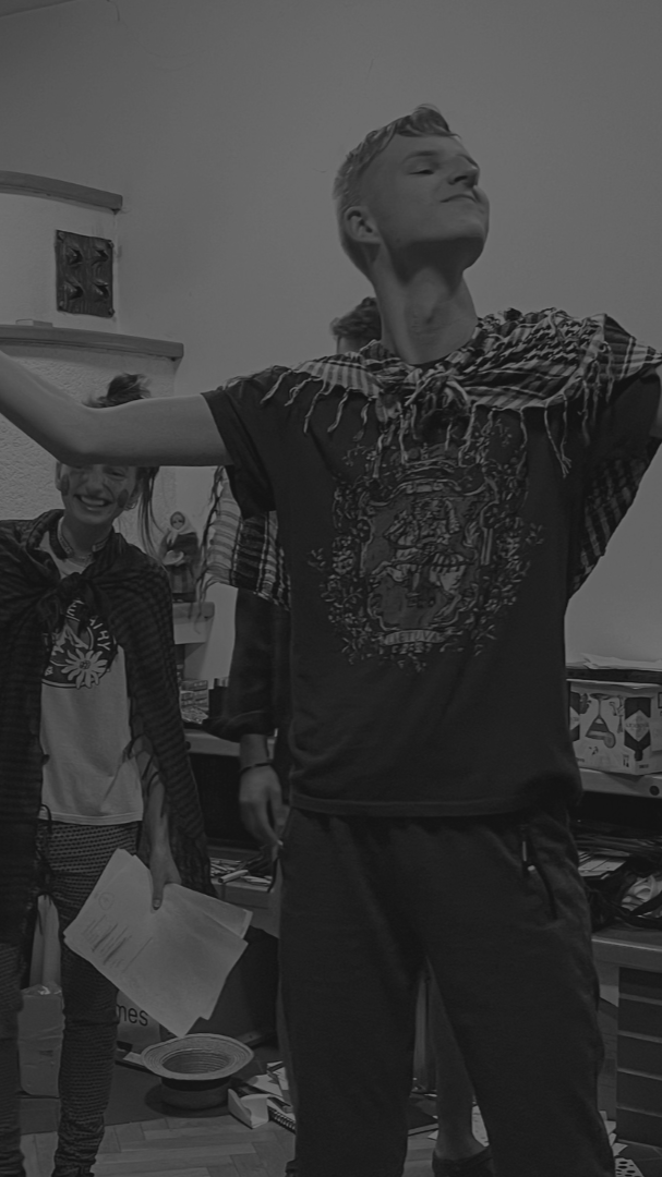 A camp participant with arms outstretched smiling in a room with other people, some holding papers.