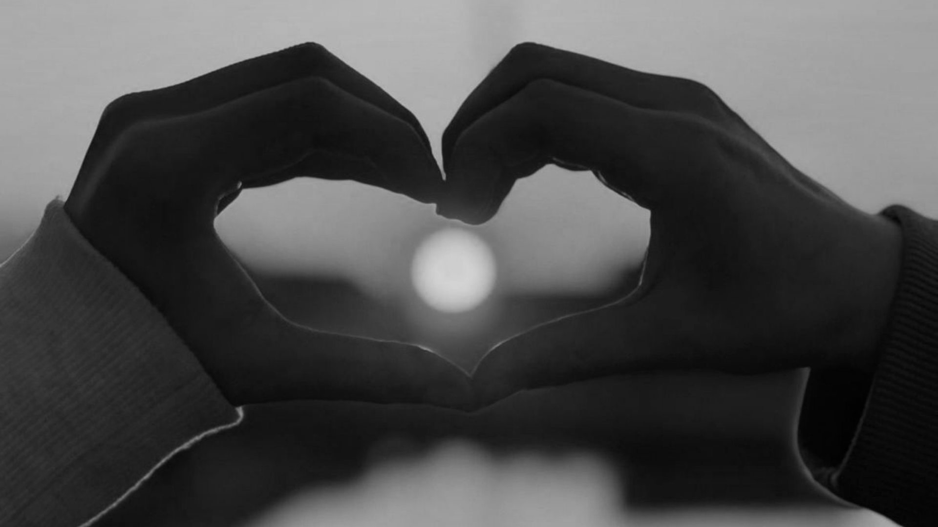 Hands forming a heart shape around the setting or rising sun in black and white.