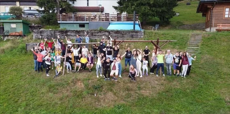 Group of people, including children and adults, gathered outdoors on a grassy hill, posing for a photo with some raising their arms and smiling.