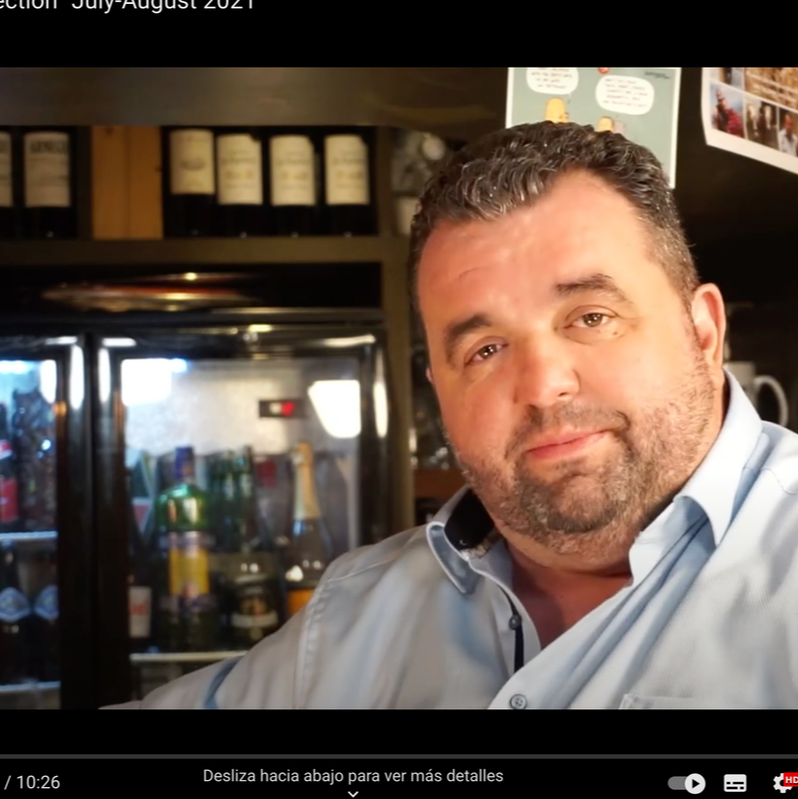 A man with dark hair and beard wearing a light blue shirt, sitting in a restaurant or bar with wine bottles and various beverages in the background.